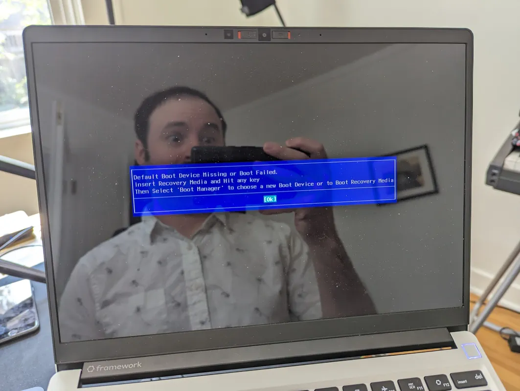 A laptop with a blue BIOS screen reading: Default Boot Device Missing or Boot Failed. The photographer is making a silly surprised face in the screen reflection.